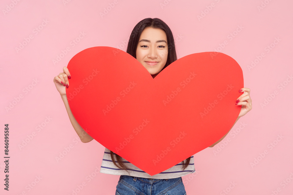 Portrait of romantic lovely woman with long brunette hair holding big red heart expressing love and gentle, wearing striped T-shirt. Indoor studio shot isolated on pink background. Portrait of romantic lovely woman with long brunette hair holding big red heart expressing love and gentle, wearing striped T-shirt. Indoor studio shot isolated on pink background.