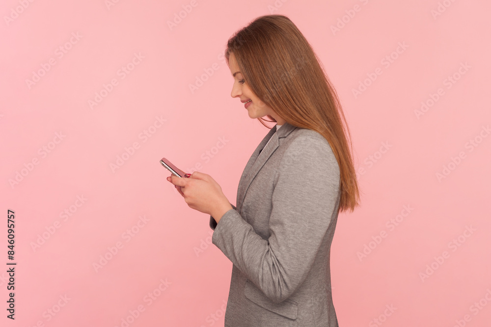 Side view portrait of smiling satisfied woman with brown hair using smart phone, chatting with friends, communicating online, wearing business suit. Indoor studio shot isolated on pink background. Side view portrait of smiling satisfied woman with brown hair using smart phone, chatting with friends, communicating online, wearing business suit. Indoor studio shot isolated on pink background.