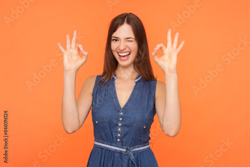 Portrait of playful positive attractive brunette woman wearing denim dress showing okay sign with both hands winking to camera. Indoor studio shot isolated on orange background.