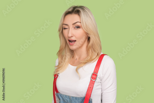 Portrait of playful optimistic adult blond woman standing winking to camera, flirting with handsome man, wearing denim overalls. Indoor studio shot isolated on light green background