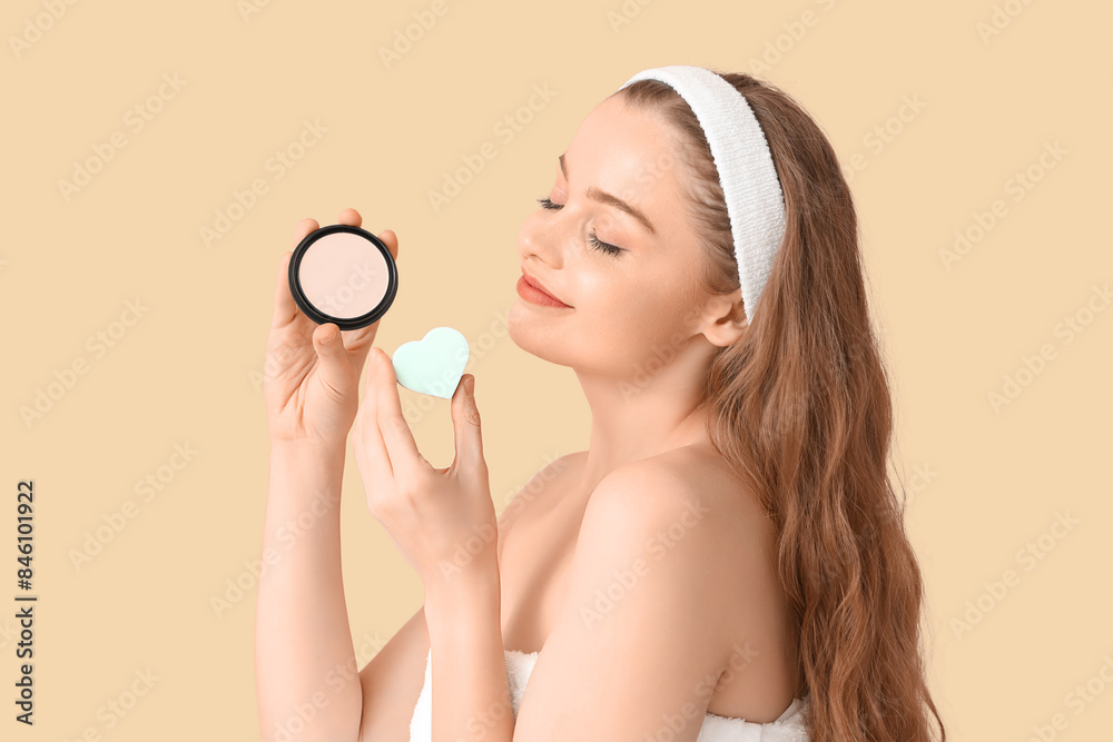 Beautiful young woman with heart-shaped sponge and makeup powder on beige background