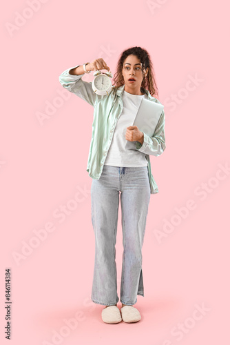Hurrying young African-American woman with laptop and alarm clock on pink background