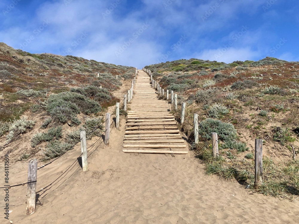 Rustic wooden staircase ascends through a sandy, shrub-covered hill at Baker Beach in San Francisco.