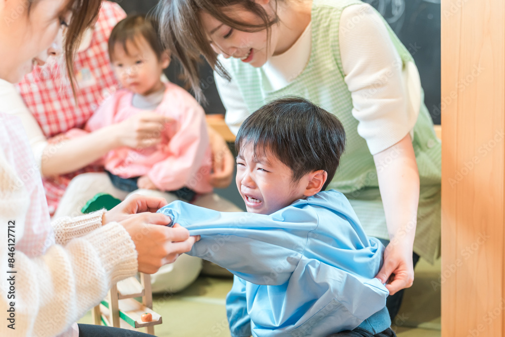 Obraz premium 保育園・幼稚園でお着替えをする保育士の女性とぐずるイヤイヤ期の園児の男の子 