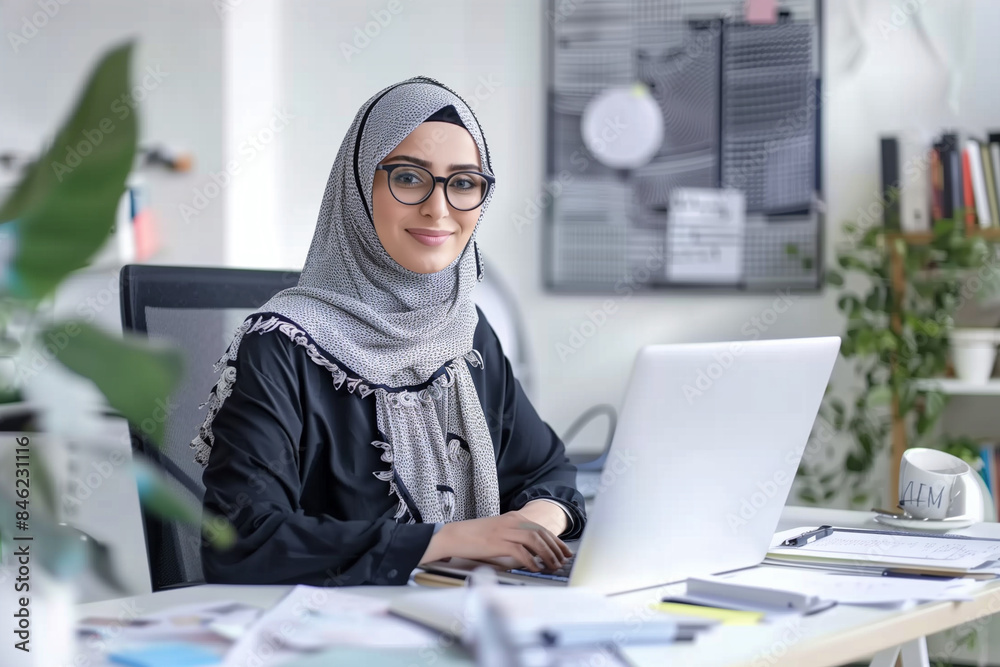 A woman wearing a black scarf and glasses is sitting at a desk with a laptop. She is focused on her work A woman wearing a black scarf and glasses is sitting at a desk with a laptop. She is focused on her work