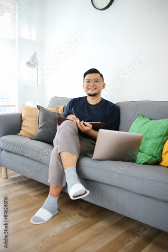 Asian man working from home using laptop and taking notes in a cozy living room © Gatot
