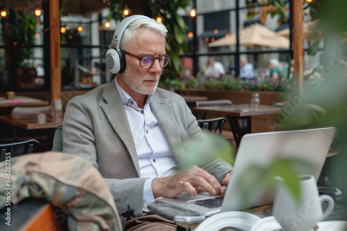 Wallpaper Mural Focused elderly man with headphones working on a laptop, seated at an outdoor cafe, exuding concentration and dedication in a bustling urban environment Torontodigital.ca