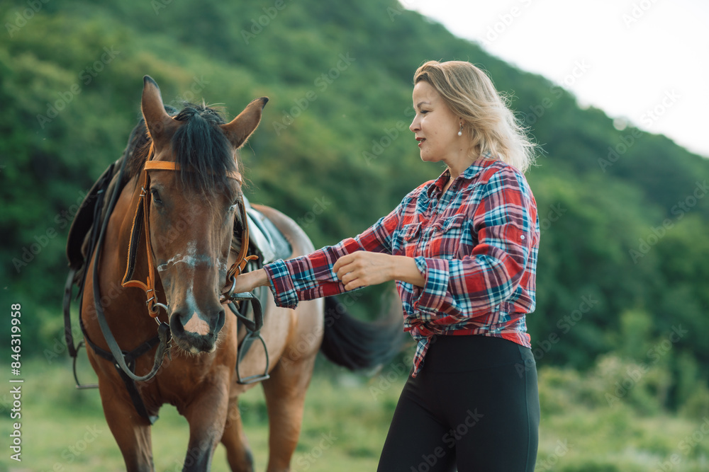 Obraz premium Happy blonde with horse in forest. Woman and a horse walking through the field during the day. Dressed in a plaid shirt and black leggings.