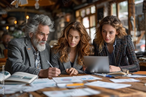 A senior man and two young women work on a business plan, reviewing papers and using a laptop while seated at a table in a cafe