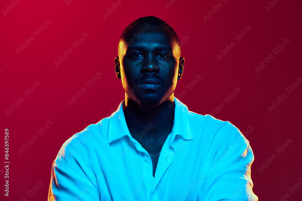 Portrait of African-American man in casual white shirt posing with serious face, showing confidence against red background in neon light. Concept of human emotions facial expression, fashion, business Portrait of African-American man in casual white shirt posing with serious face, showing confidence against red background in neon light. Concept of human emotions facial expression, fashion, business