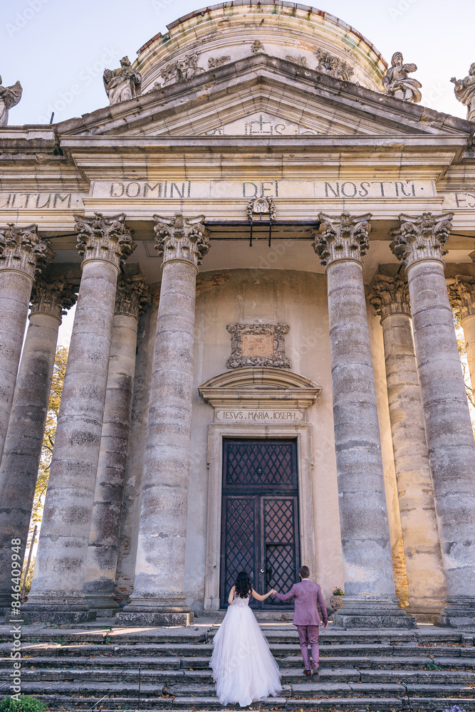 Obraz premium a beautiful couple of newlyweds are holding hands and walking towards an old building with columns and beautiful big doors.