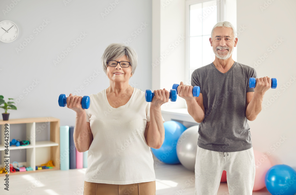 Happy sporty senior mature people man and woman wearing sportswear looking at camera and doing fit exercises with dumbbells in gym together. Concept of sport and fitness in retirement. Happy sporty senior mature people man and woman wearing sportswear looking at camera and doing fit exercises with dumbbells in gym together. Concept of sport and fitness in retirement.