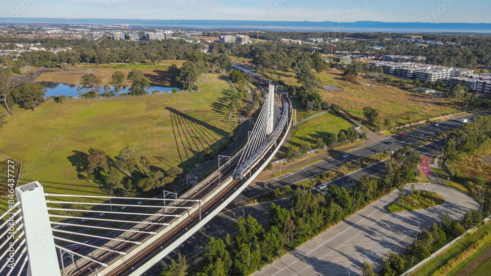 Fototapeta premium Aerial drone view of a train crossing the railway bridge as it travels toward Rouse Hill Station on the metro northwest railway line, Greater Sydney, NSW Australia in June 2024