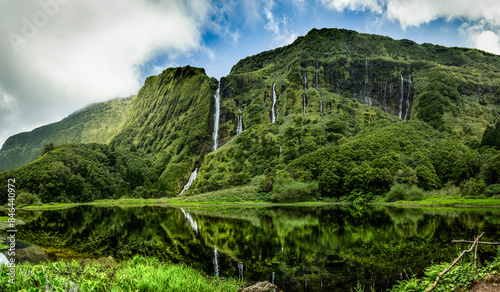 A stunning landscape of Flores Island in the Azores, a serene lake surrounded by lush mountains and cascading waterfalls. The vibrant scenery captures the untouched beauty of this volcanic paradise.