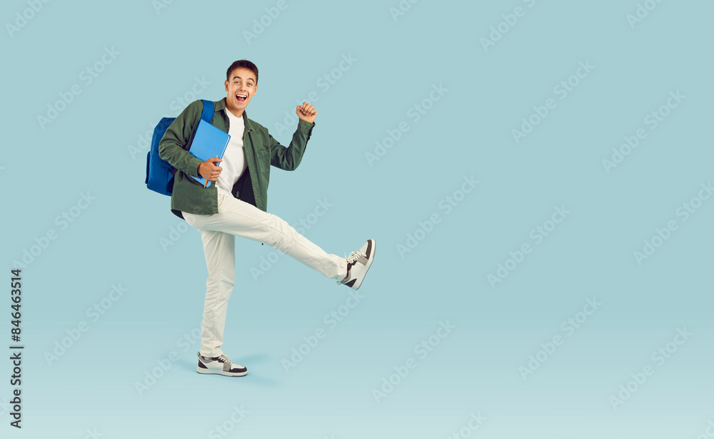Happy cheerful university, college or high school student boy in casual clothes with backpack and notebook walking in funny way on light blue blank copy space studio background Happy cheerful university, college or high school student boy in casual clothes with backpack and notebook walking in funny way on light blue blank copy space studio background
