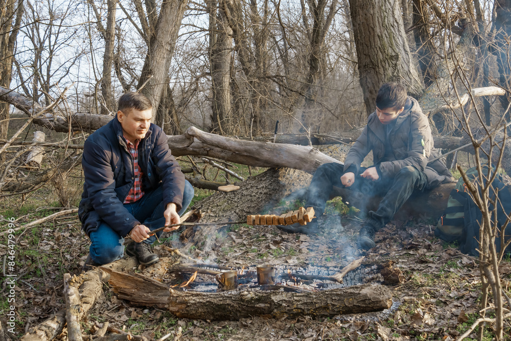 Two men make a campfire for cooking in the forest, father and son sitting on a log while hiking and outdoor activities, early spring landscape