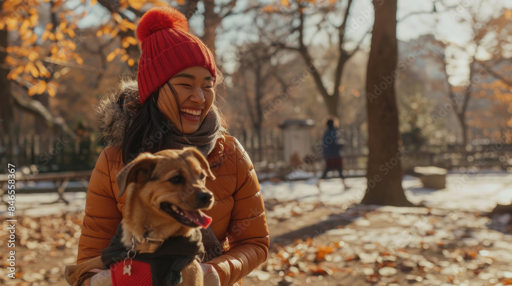 A woman in a red beanie and brown jacket smiles while holding her brown dog in a park setting, during autumn