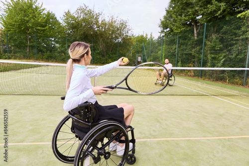 Woman in wheelchair playing tennis on outdoor court, focusing on ball with opponent in background © Robert Kneschke
