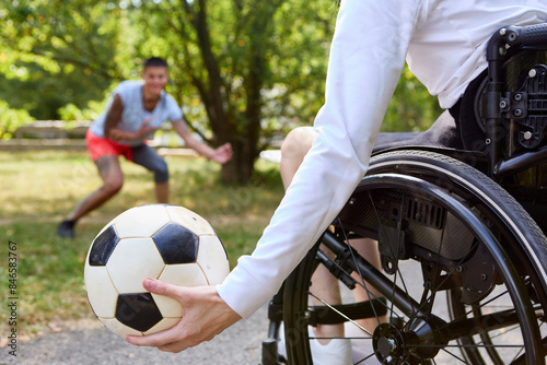 Fototapeta Naklejka Na Ścianę i Meble -  Person in wheelchair holding a soccer ball, playing with friend at the park