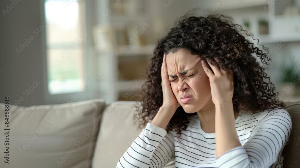 An American woman with curly hair and holding her head in pain while sitting on the sofa at home An American woman with curly hair and holding her head in pain while sitting on the sofa at home