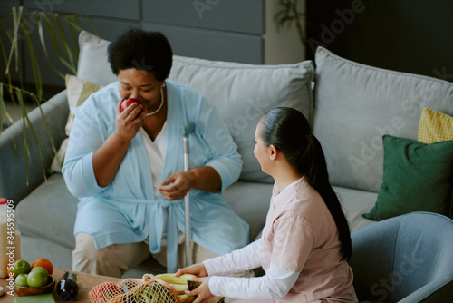 Mature patient inhaling fresh and sweet apple scent while young nurse dressed in scrubs sitting next to her keeping unpacking grocery products