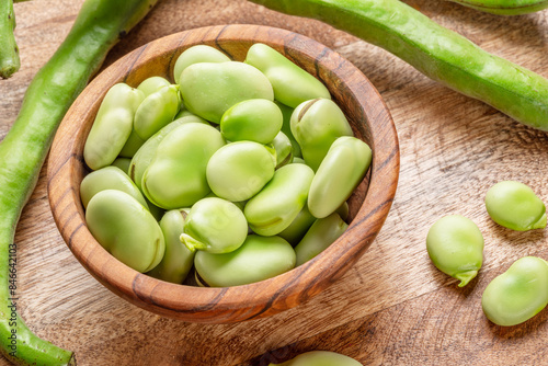 Broad beans in wooden bowl and broad bean pods near it on wooden table. Top view.