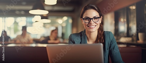 A joyful businesswoman, wearing glasses, types on a laptop inside an office, looking pleased with her accomplishments, with a backdrop of an office environment with a copy space image available.