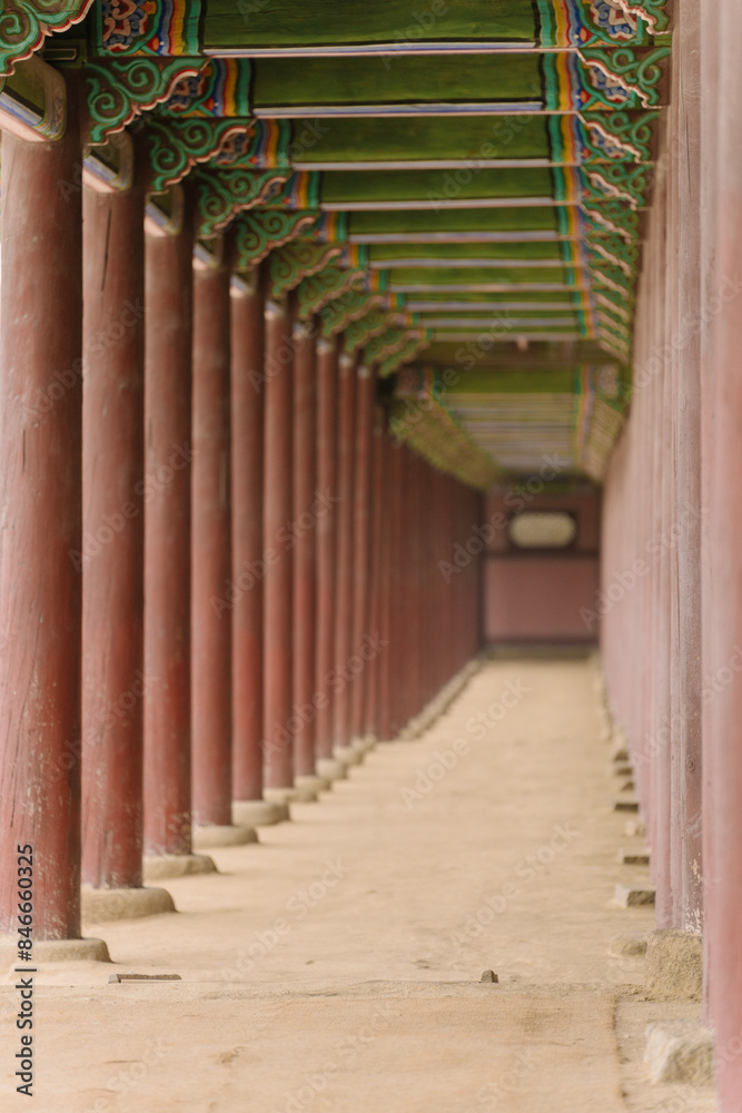 Fototapeta premium a long corridor with traditional Korean architecture, featuring colorful wooden beams and a series of red pillars leading to a vanishing point.