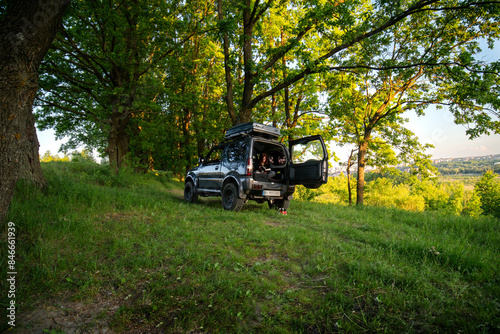 A young woman sits in the trunk of an SUV and reads a book. Off road vehicle outdoors. Summer green trees and grass, Forest. Camping. Copy space