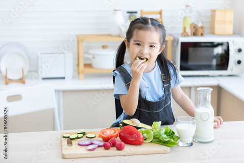 cute asian little child girl eating slice a cucumber with different vegetables in the kitchen