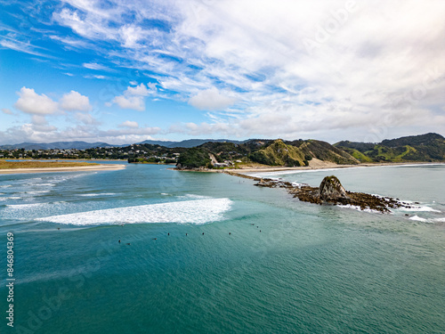 Aerial view of surfers waiting for waves in prime spot by rocky peninsular in Mangawhai beach, New Zealand. 