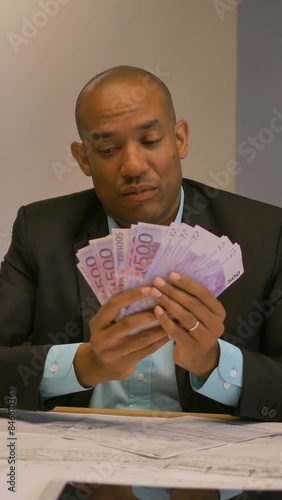 Young male entrepreneur counting money at his desk. Portrait. Looking at camera. He hold it like fan. Vertical shot.