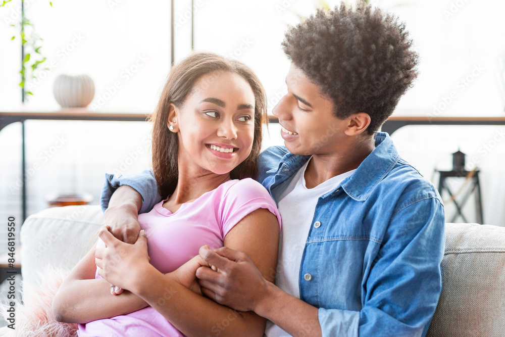 African American teen couple is sitting on a couch in a living room. The guy is holding the girls hand, and they are looking at each other with smiles on their faces African American teen couple is sitting on a couch in a living room. The guy is holding the girls hand, and they are looking at each other with smiles on their faces