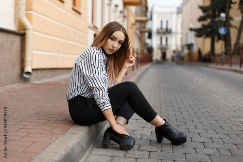 Fashionable beautiful girl hipster model with nose piercing in a fashion striped shirt with jeans with boots sits in the city