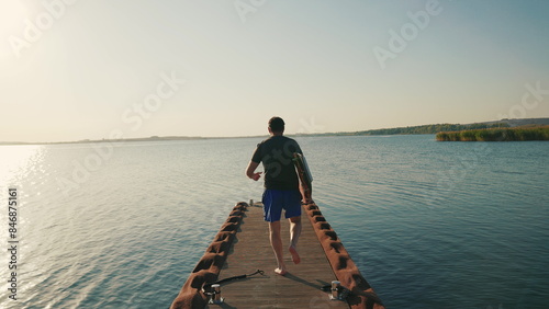 A male surfer walks on a pier on a lake against the sun, holding a water board in his hands. A male surfer walks along the pier with a small surfboard in his hands. Summer water sports