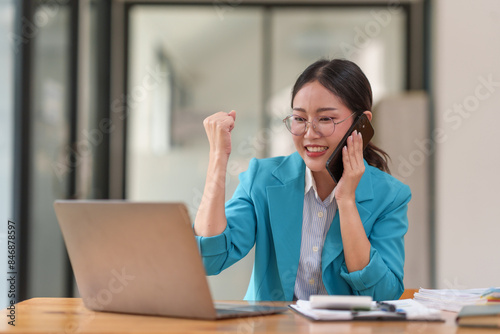 Businesswoman celebrating success while on phone call at laptop in office. Happy and motivated, achieving goals. Professional workspace.