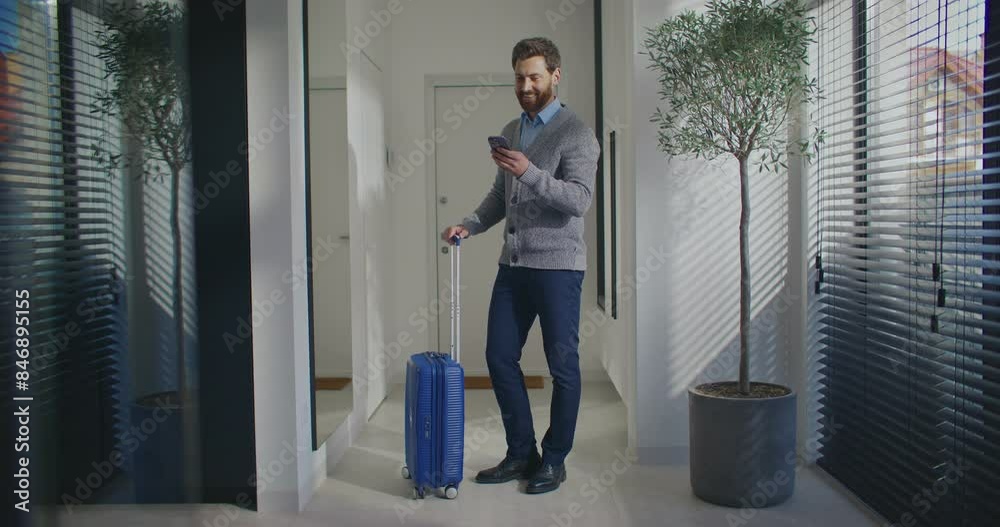 Bearded Caucasian man standing in middle of foyer. Holding handle of suitcase with one hand. Looking outside. Making plans. Waiting for taxi. Chatting with friend or family on smartphone. Bearded Caucasian man standing in middle of foyer. Holding handle of suitcase with one hand. Looking outside. Making plans. Waiting for taxi. Chatting with friend or family on smartphone.