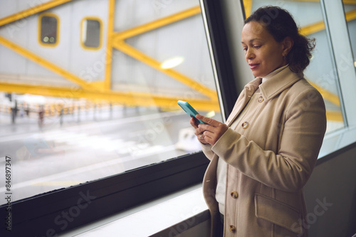 Woman in a beige coat using a smartphone by a window at an airport