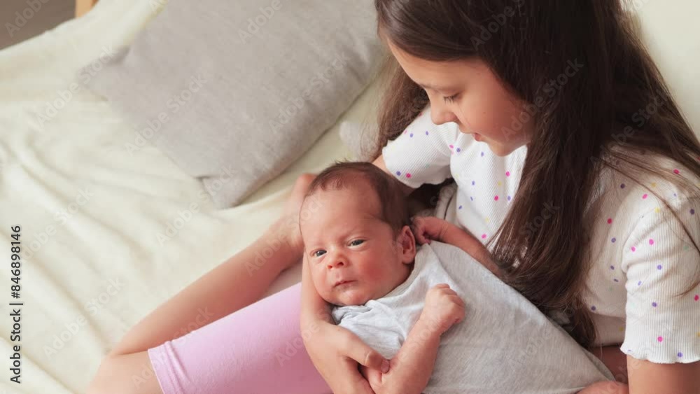Cute older little girl sitting on the sofa holding her newborn brother in her arms while kid falling asleep rocking the baby with love Cute older little girl sitting on the sofa holding her newborn brother in her arms while kid falling asleep rocking the baby with love