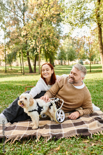 A man and woman in casual attire sit on a blanket with their dog in a peaceful park setting. © LIGHTFIELD STUDIOS