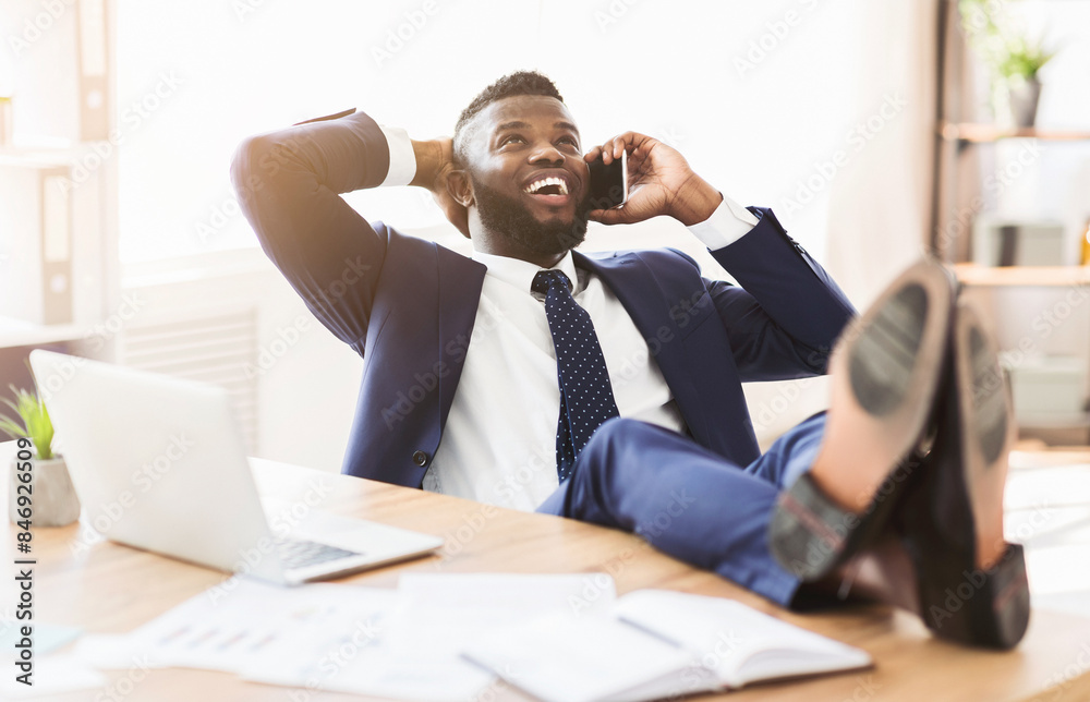 Cheerful african manager talking by phone while having break at office, leaning back on chair and putting legs on table, panorama with copy space Cheerful african manager talking by phone while having break at office, leaning back on chair and putting legs on table, panorama with copy space