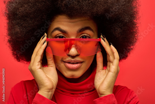 A stylish African American woman with curly hairdohairstyle holds up a pair of red glasses.