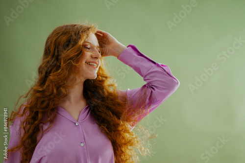 Beautiful happy smiling redhead freckled woman with long curly hair wearing purple satin blouse, posing on green background. Close up studio portrait. Copy, empty, blank space for text
