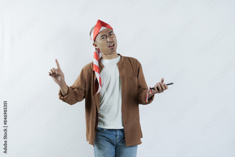 Asian man holding cell phone and showing gesture of disappointment expression. Bald-headed Asian man wearing a headband typical of the independence day of the republic of indonesia