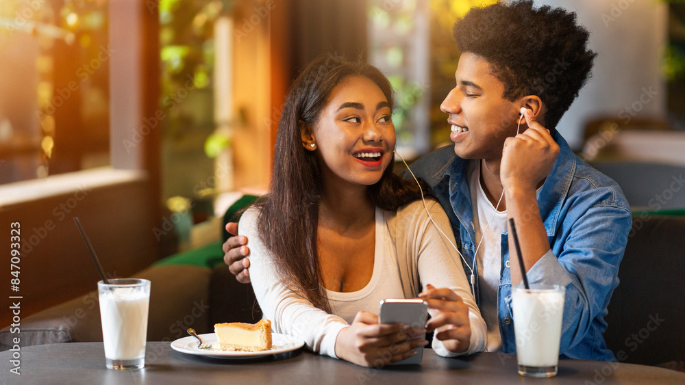 A young black couple enjoys sharing earphones and listening to music while sitting at a cafe table, smiling and laughing together on a sunny day. Each has a glass of milk and a slice of cake A young black couple enjoys sharing earphones and listening to music while sitting at a cafe table, smiling and laughing together on a sunny day. Each has a glass of milk and a slice of cake