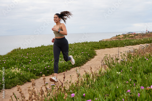 Hispanic female runner running along a dirt path with the ocean in the background, wearing dark tights and olive top.