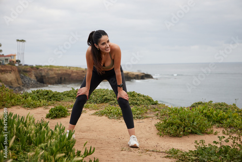 Female hispanic runner resting arms on her legs, wearing black leggings and a olive top, ocean view in the background.