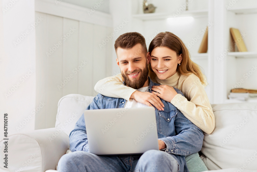 Smiling couple sits on the couch, looking at a laptop in a cozy home setting, showing affection and joyful companionship. Smiling couple sits on the couch, looking at a laptop in a cozy home setting, showing affection and joyful companionship.