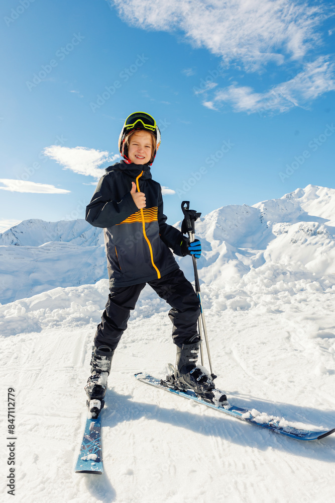 Active adorable teenager caucasian kid boy portrait with ski in helmet, goggles and bright suit enjoy winter extreme sport activities. Little child skiing luxury alpine resort in mountains thumbs up Active adorable teenager caucasian kid boy portrait with ski in helmet, goggles and bright suit enjoy winter extreme sport activities. Little child skiing luxury alpine resort in mountains thumbs up