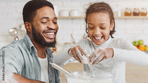 Cute african girl adding eggs into dough bowl, father teaching daughter cooking in kitchen